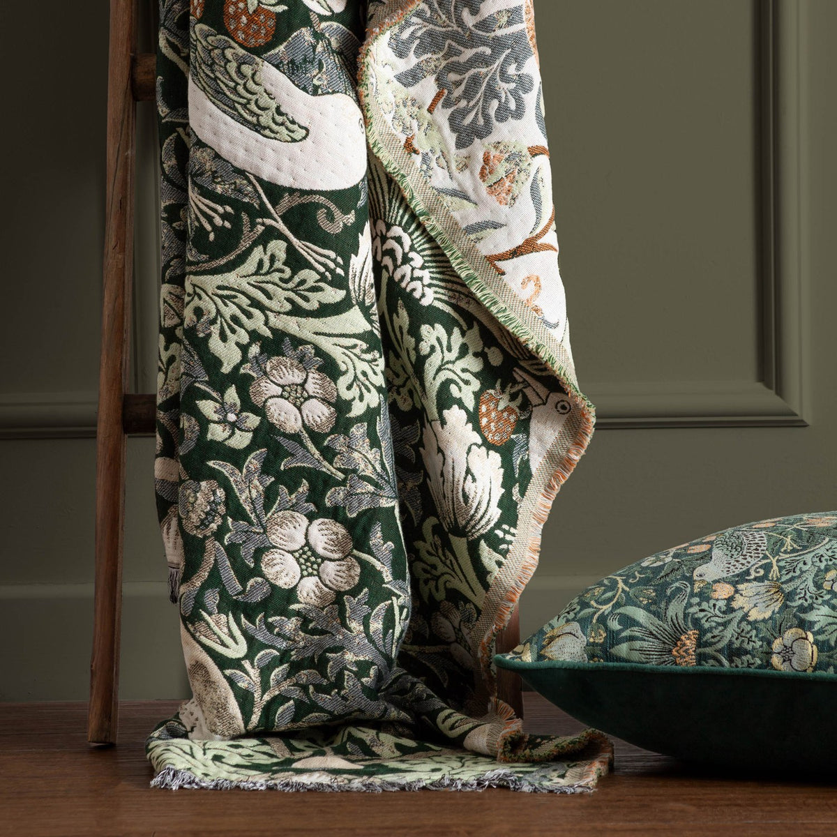 Strawberry Thief Jacquard Cotton Throw in forest green, draped over a wooden ladder showing bird and floral pattern with cream reverse, beside a matching cushion.