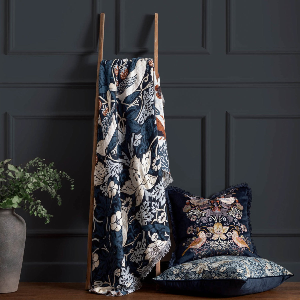 Strawberry Thief Jacquard Cotton Throw in indigo draped over a wooden ladder, showing bird and floral pattern with fringed edge, styled beside matching cushions in a dark-panelled room.