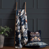 Strawberry Thief Jacquard Cotton Throw in indigo draped over a wooden ladder, showing bird and floral pattern with fringed edge, styled beside matching cushions in a dark-panelled room.