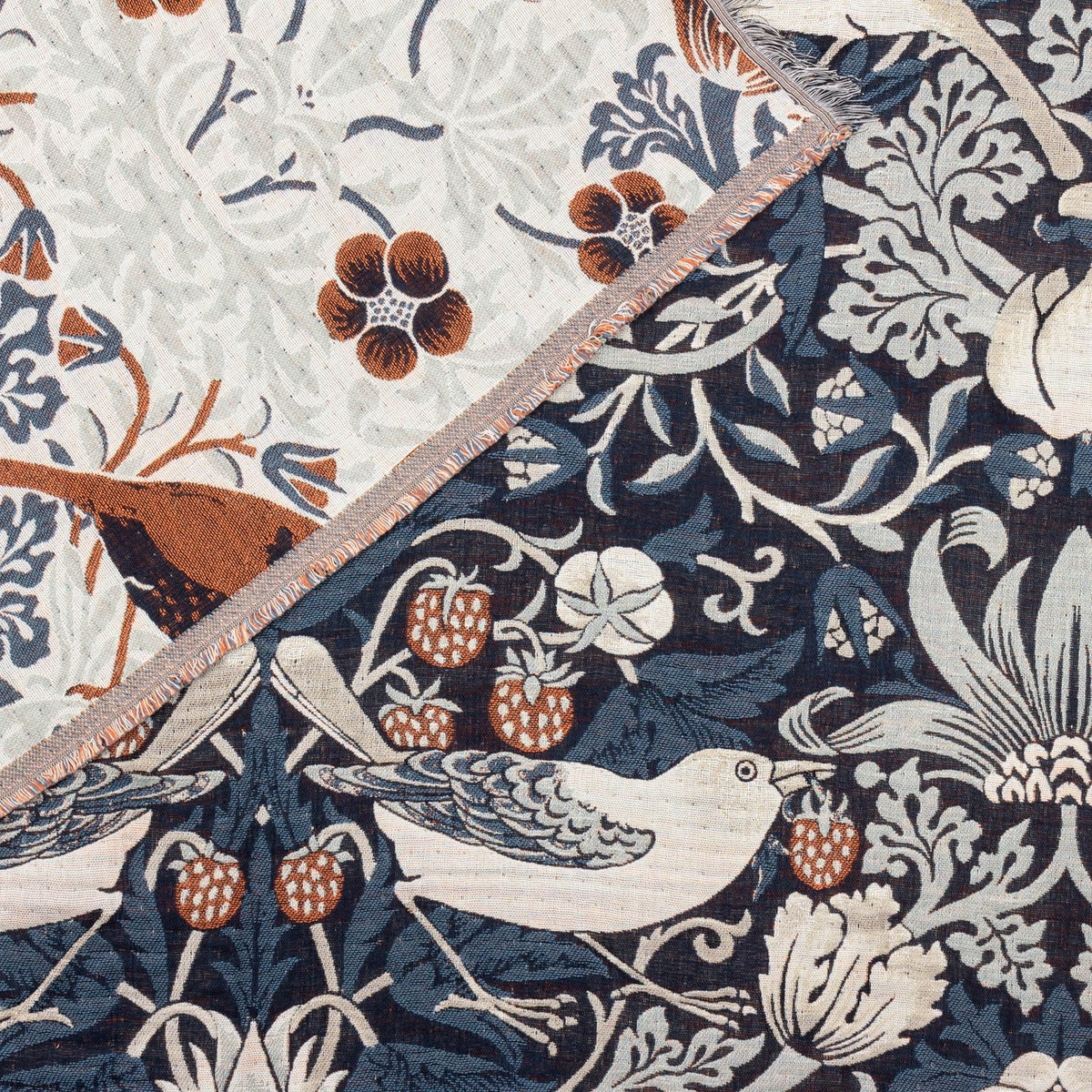 Strawberry Thief Jacquard Cotton Throw in indigo, close-up of reversible woven fabric showing birds, strawberries and florals in cream, rust and blue tones with fringed edge trim.