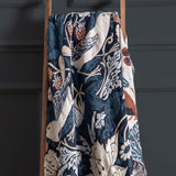 Strawberry Thief Jacquard Cotton Throw in indigo, draped over a wooden ladder, showing a woven bird, strawberry and floral pattern in blue, cream and rust with fringed edges.