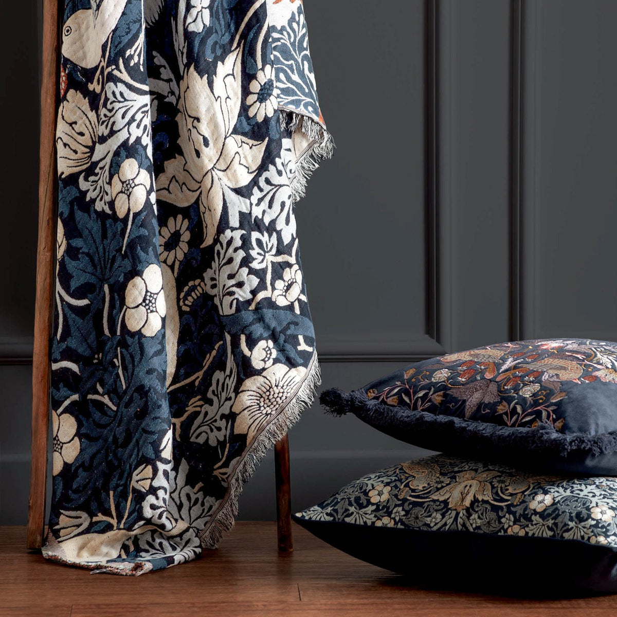 Strawberry Thief Jacquard Cotton Throw in indigo, draped over a wooden chair with fringed edge, shown beside matching cushions in a dark-panelled room.