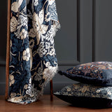 Strawberry Thief Jacquard Cotton Throw in indigo, draped over a wooden chair with fringed edge, shown beside matching cushions in a dark-panelled room.
