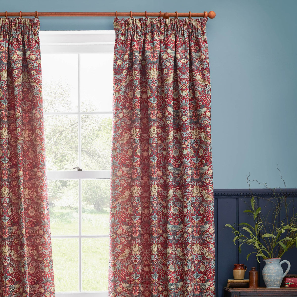 Strawberry Thief pencil pleat curtains in berry red jacquard fabric with bird and floral pattern, hung by a window in a light blue room.