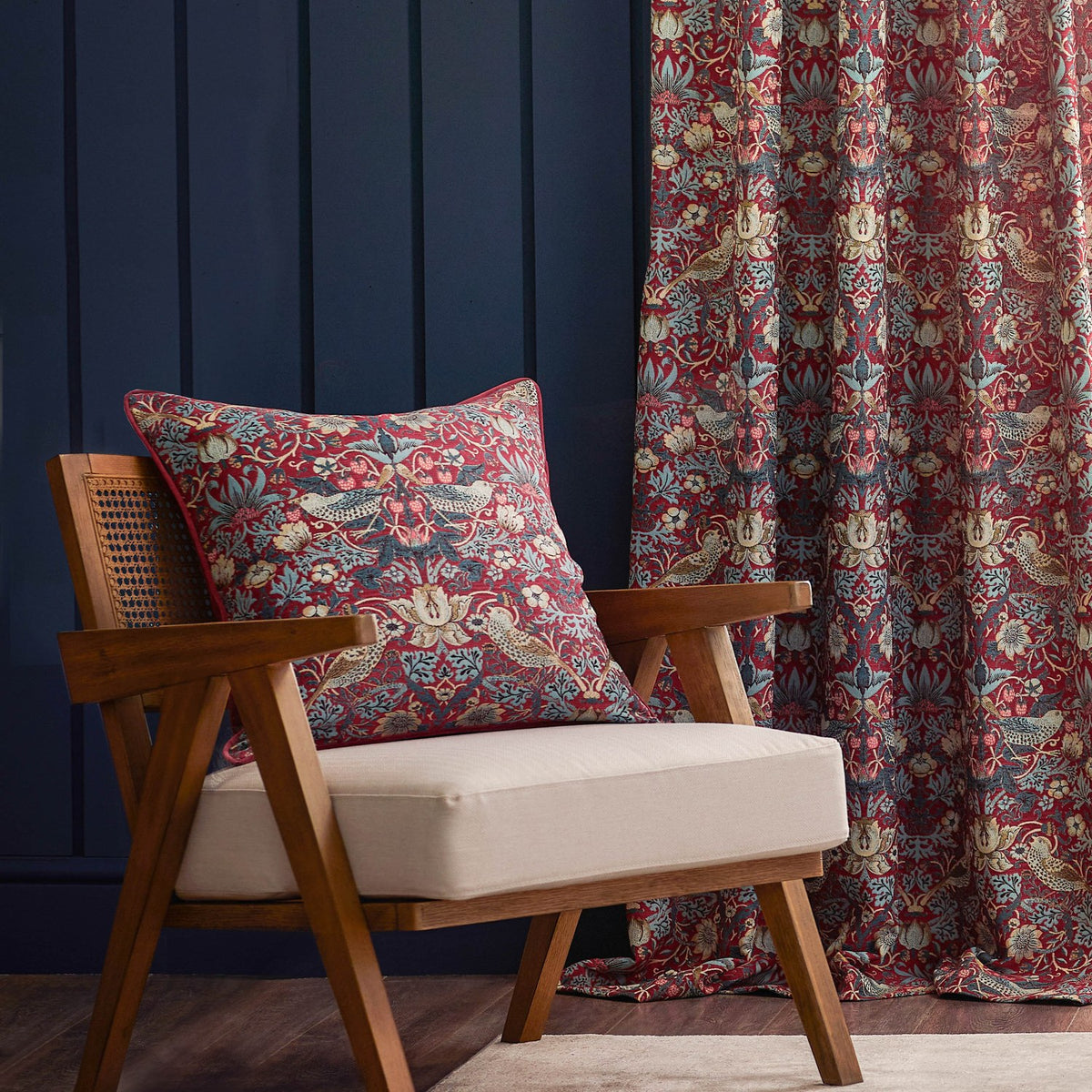 Strawberry Thief pencil pleat curtain in berry red jacquard fabric with bird and floral pattern, shown beside a matching cushion on a wooden armchair against a dark blue panelled wall.