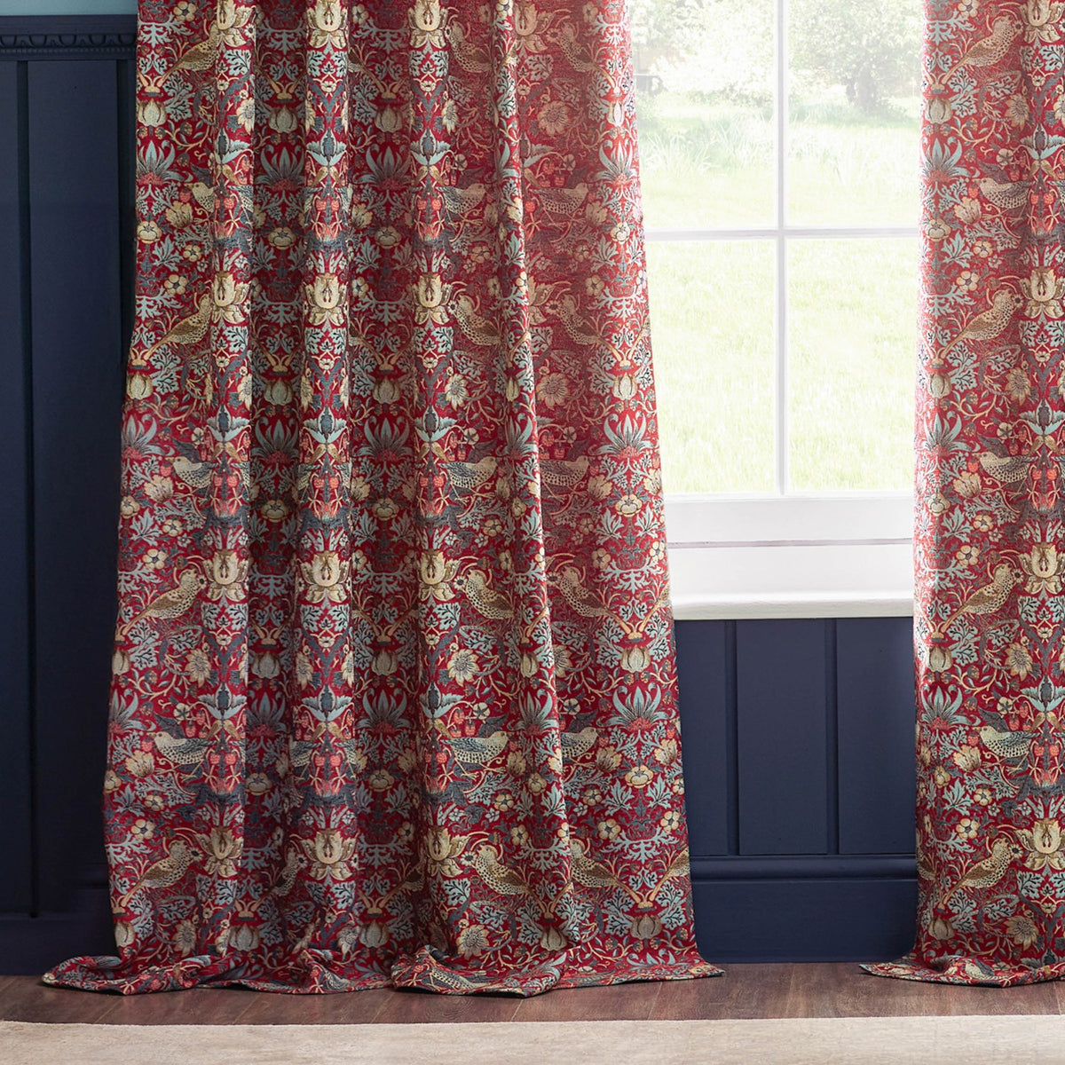 Strawberry Thief pencil pleat curtains in berry red jacquard fabric with bird and floral pattern, hung pleated at a window beside navy panelled walls.