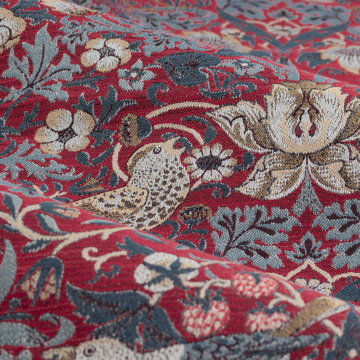 Close-up of Strawberry Thief pencil pleat curtain fabric in berry red, showing a jacquard-woven bird, floral, and strawberry motif in blue-grey and cream tones.