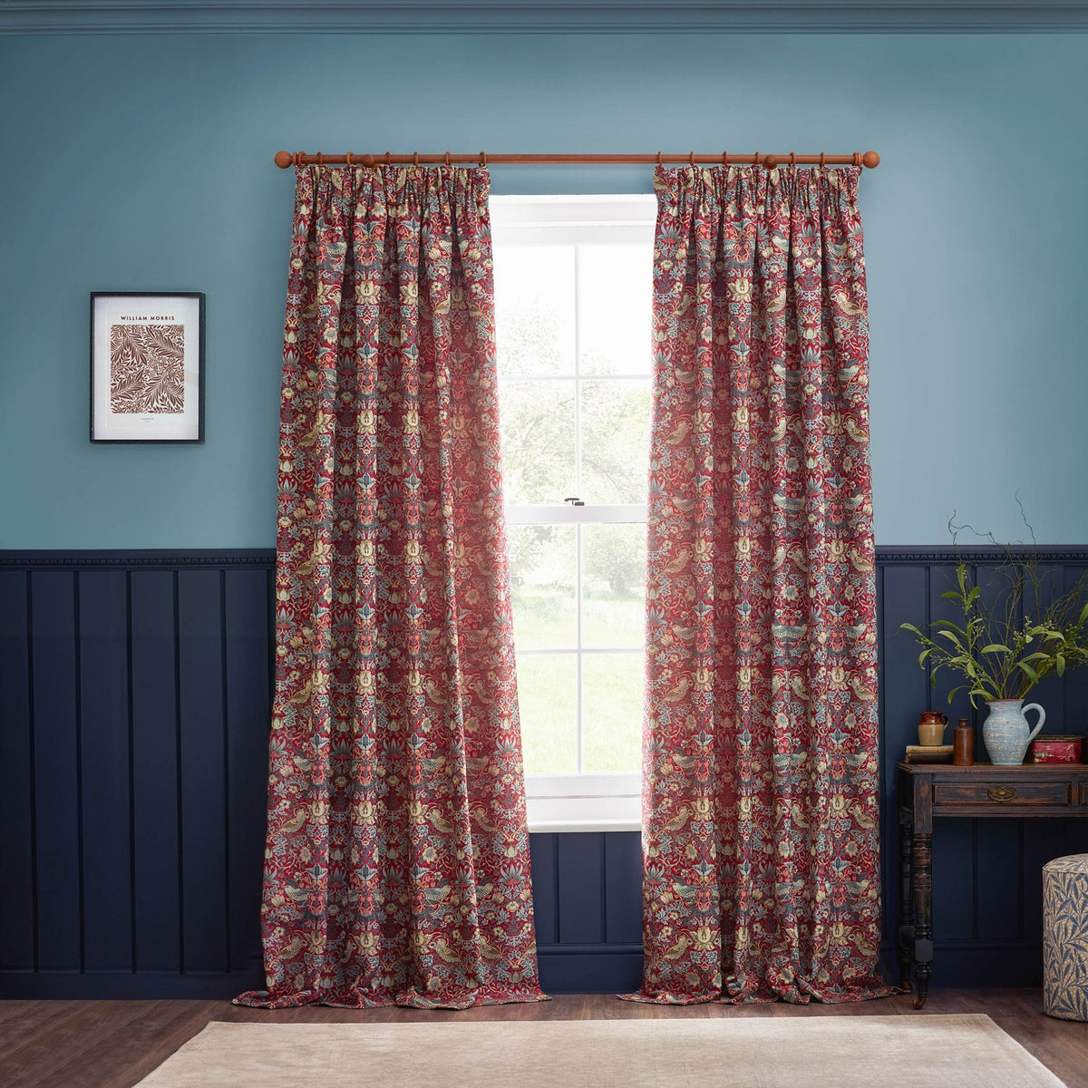 Strawberry Thief jacquard pencil pleat curtains in berry red hung from a wooden pole either side of a window in a blue and navy panelled living room.