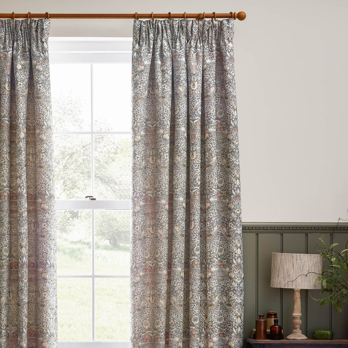 Strawberry Thief pencil pleat curtains in biscuit, jacquard-woven bird and foliage pattern in grey, cream and brown, shown pleated on a wooden pole beside a window.