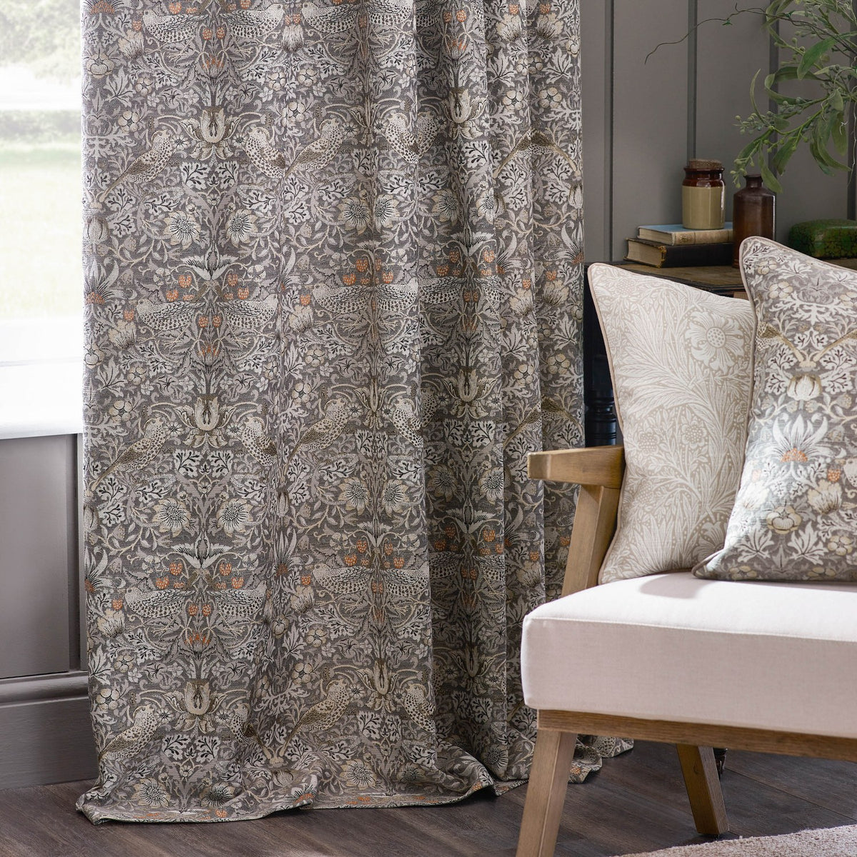 Strawberry Thief pencil pleat curtain in biscuit tone with woven bird and foliage jacquard pattern, shown hanging beside matching cushions on an armchair in a living room setting.