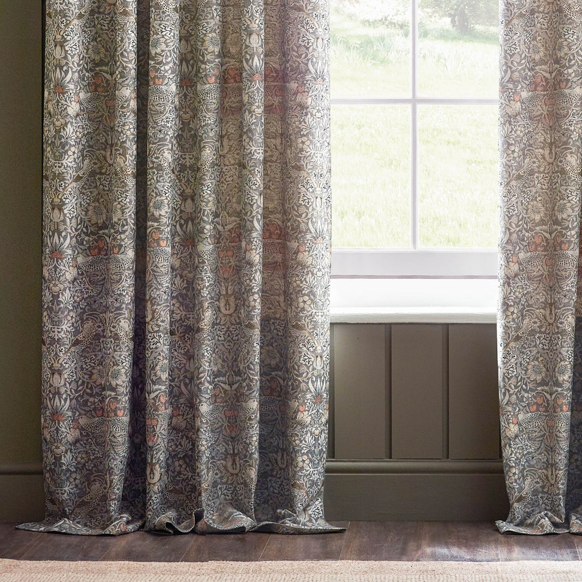 Pencil pleat curtains in Strawberry Thief jacquard, showing woven birds and foliage in grey, brown and biscuit tones, hung beside a window in a living room.