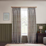 Strawberry Thief pencil pleat curtains in biscuit, jacquard-woven fabric with bird and foliage pattern, hung on a wooden pole beside a window in a living room with olive wainscoting.