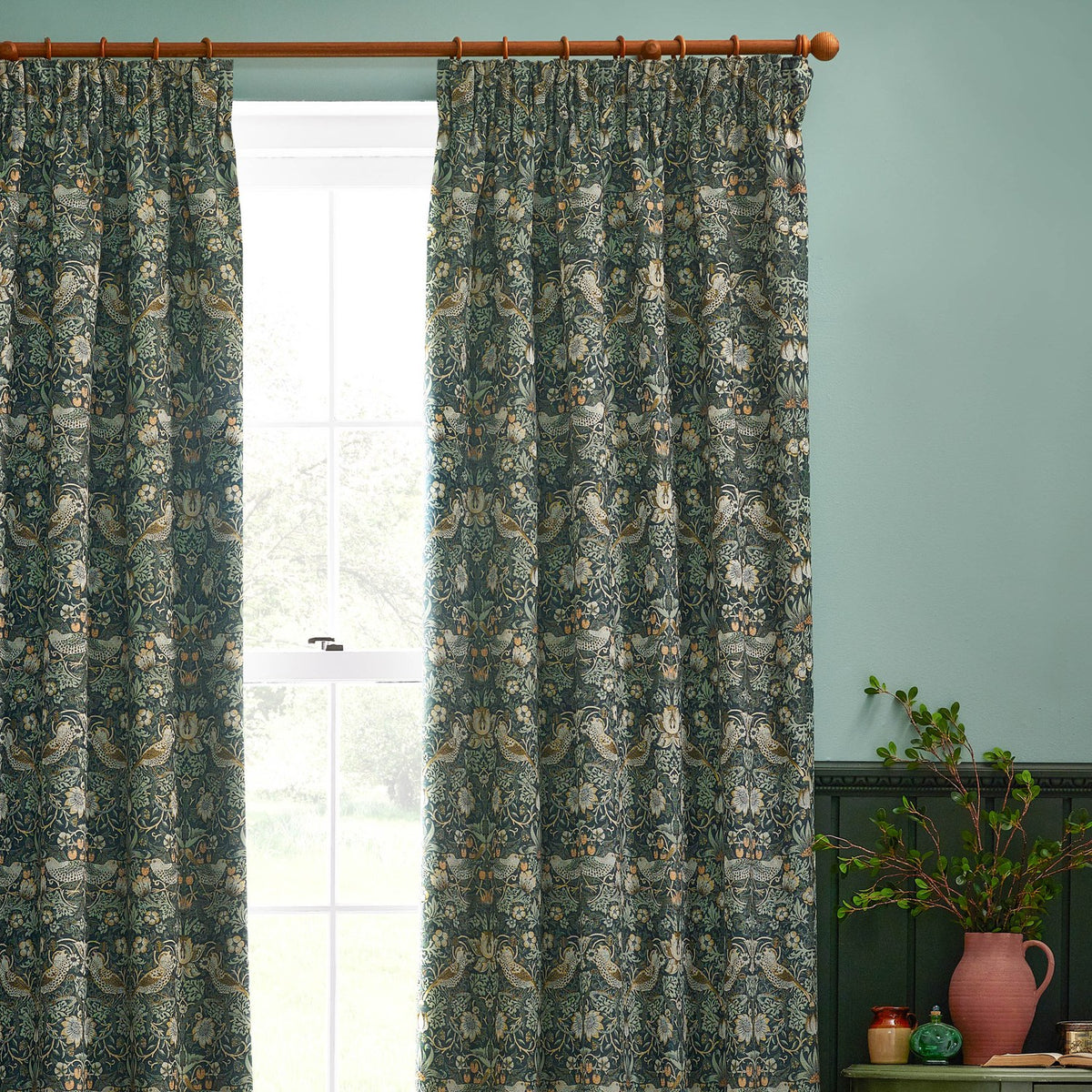 Strawberry Thief pencil pleat curtains in forest green polyester, featuring Morris & Co.'s bird and strawberry print, hung on a wooden pole beside a window in a duck-egg blue room.