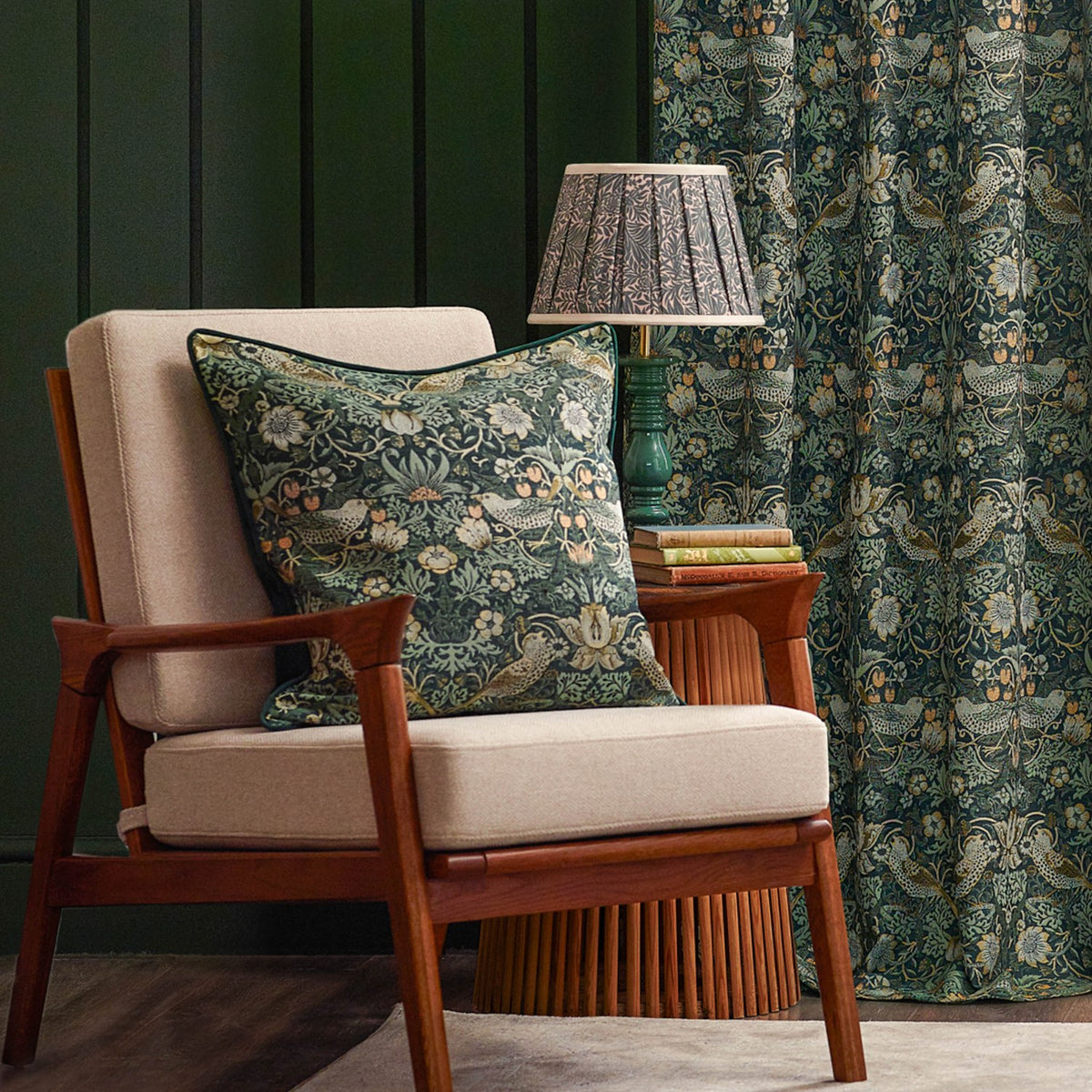 Strawberry Thief pencil pleat curtain in forest green polyester with bird and strawberry print, shown beside a matching cushion, wooden armchair, and patterned lampshade.