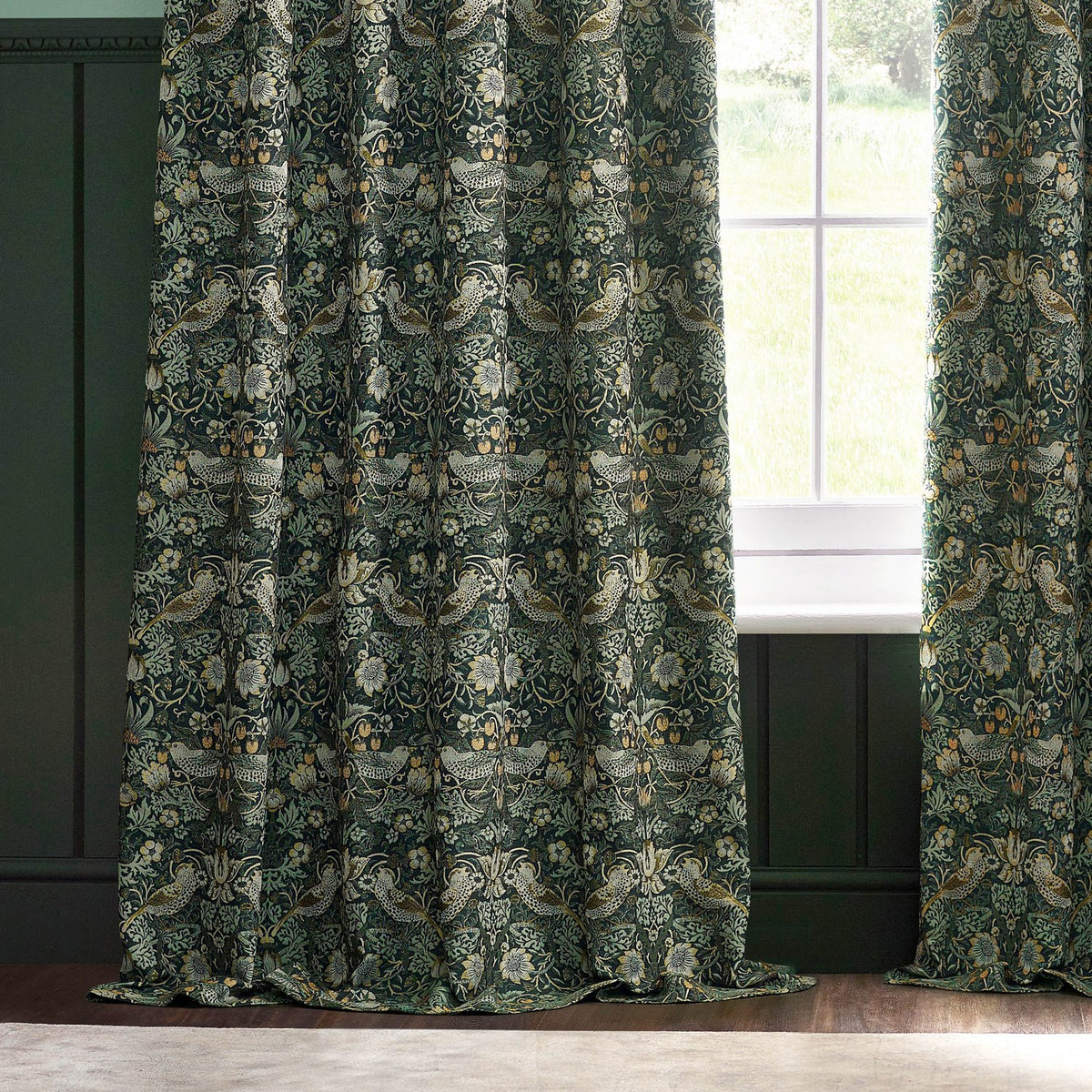 Pencil pleat curtains in the Strawberry Thief design, forest green polyester fabric with bird and floral pattern, shown hanging beside a window in a green-panelled room.