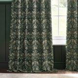 Pencil pleat curtains in the Strawberry Thief design, forest green polyester fabric with bird and floral pattern, shown hanging beside a window in a green-panelled room.