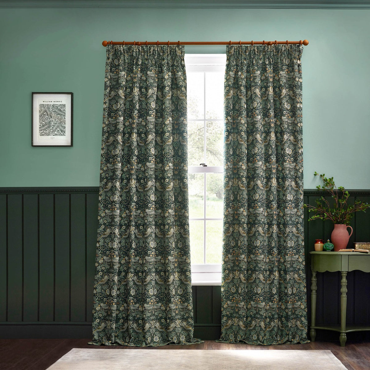 Pair of pencil pleat curtains in the Strawberry Thief design, forest green polyester fabric with bird and strawberry motif, hung on a wooden pole in a green-walled room.