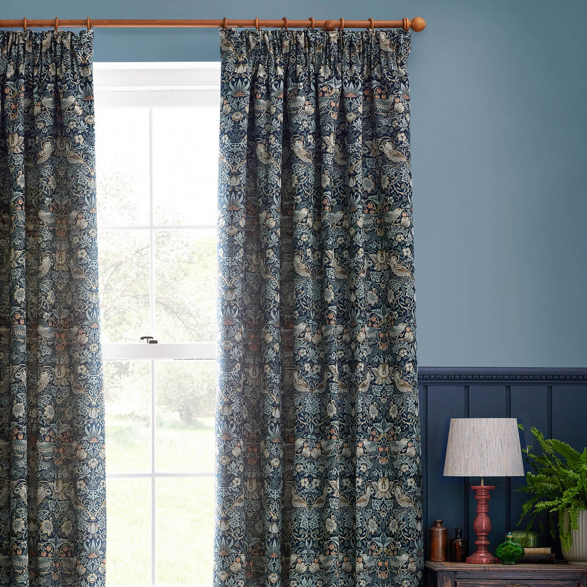 Strawberry Thief pencil pleat curtains in indigo blue jacquard fabric with a woven bird and strawberry pattern, shown hanging beside a window in a blue-walled living room.