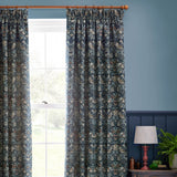 Strawberry Thief pencil pleat curtains in indigo blue jacquard fabric with a woven bird and strawberry pattern, shown hanging beside a window in a blue-walled living room.