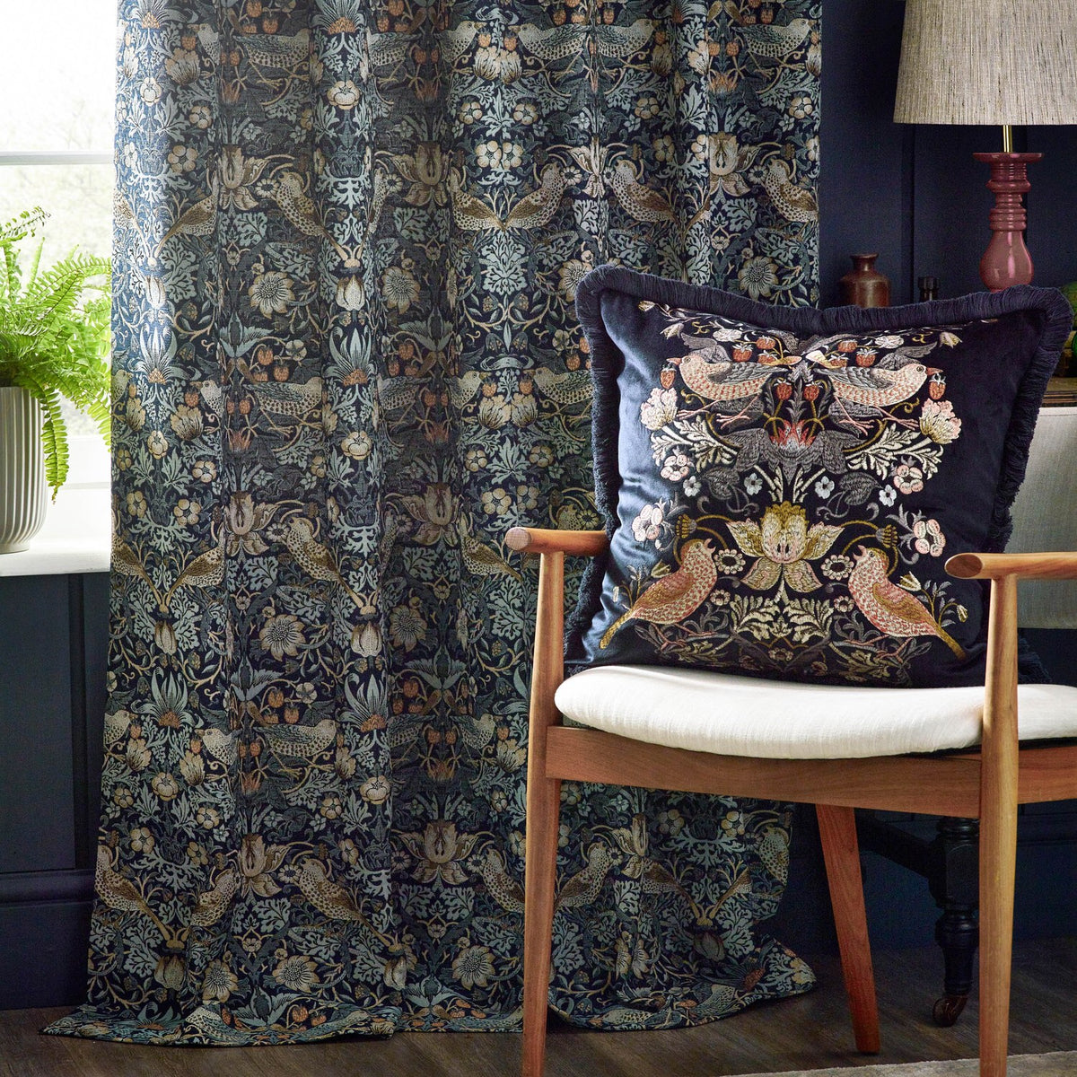 Strawberry Thief pencil pleat curtain in indigo jacquard fabric with bird and floral pattern, styled beside a matching cushion on a wooden armchair.