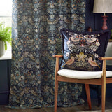 Strawberry Thief pencil pleat curtain in indigo jacquard fabric with bird and floral pattern, styled beside a matching cushion on a wooden armchair.