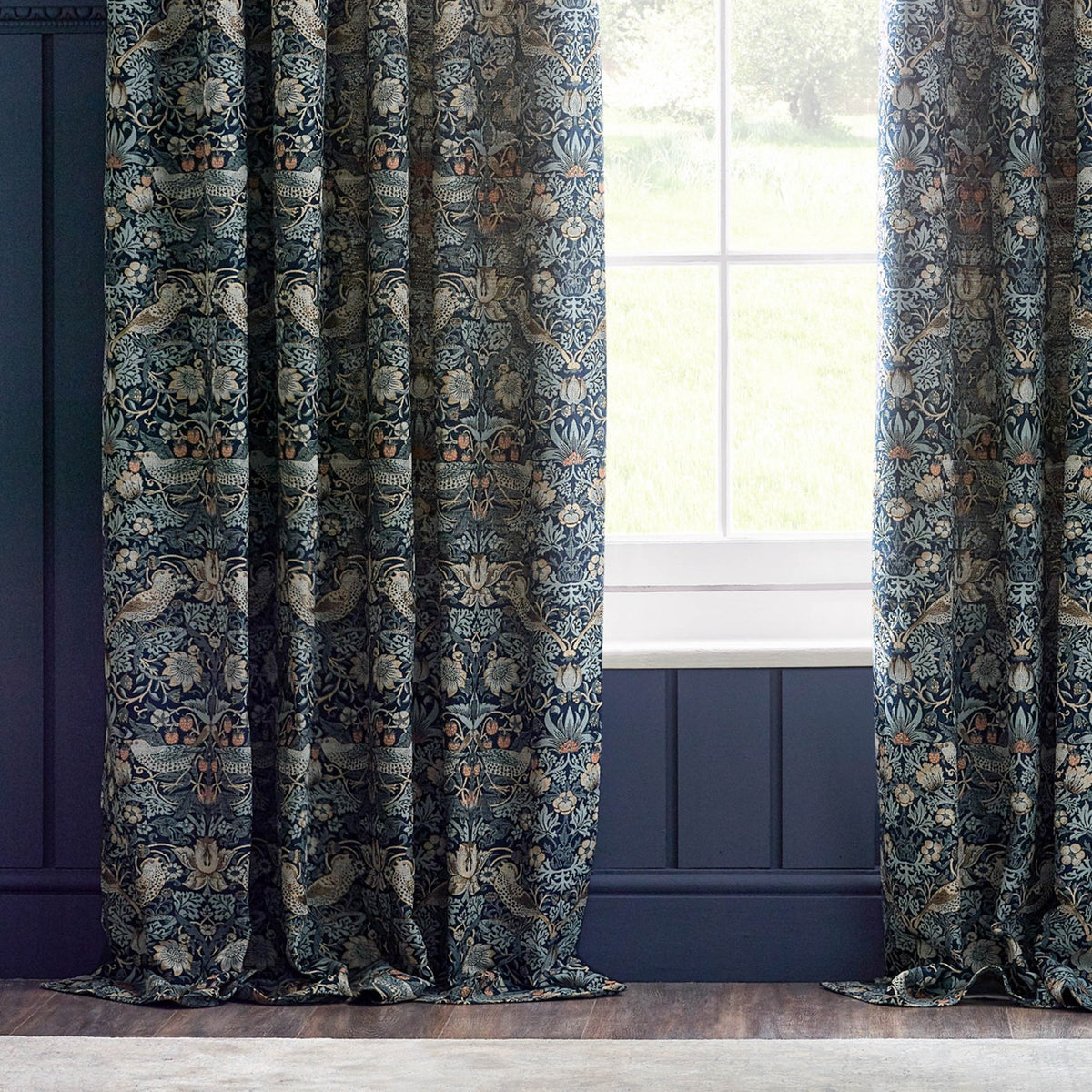Strawberry Thief pencil pleat curtains in indigo, jacquard-woven polyester with birds, strawberries and floral trails, hung beside a window in a living room setting.