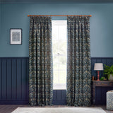 Strawberry Thief pencil pleat curtains in indigo jacquard fabric with a bird and floral pattern, shown hanging from a wooden pole in a blue-walled living room.