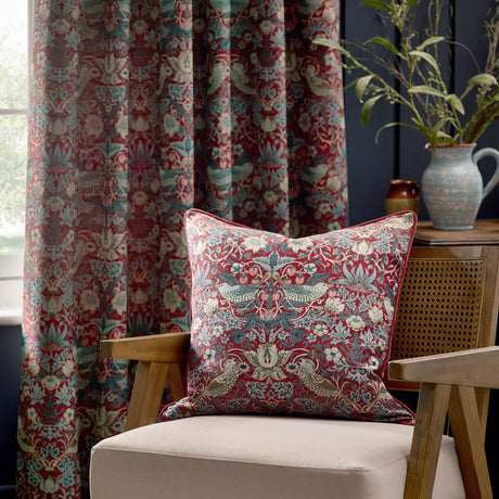 Strawberry Thief Jacquard Velvet Cushion in Berry with bird and floral pattern, styled on a beige armchair against matching curtains in a living room setting.