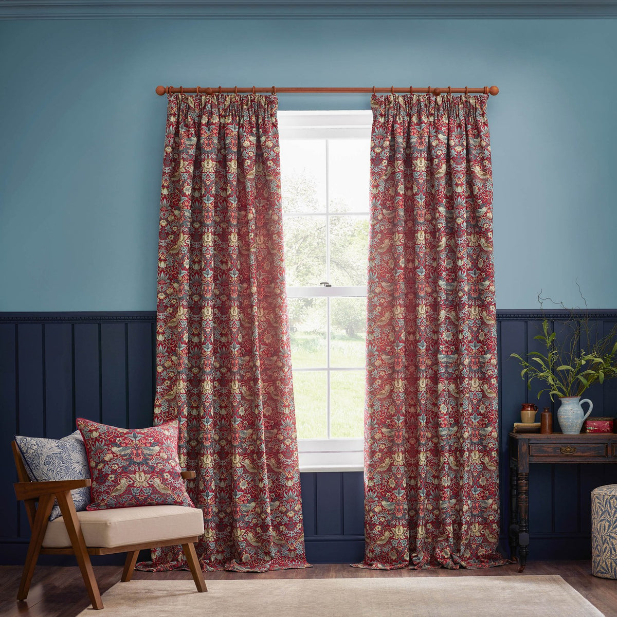 Strawberry Thief Jacquard Velvet Cushion in Berry on a beige armchair, showing a red patterned bird and floral design against a blue panelled living room wall.