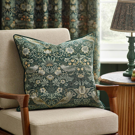 Strawberry Thief Jacquard Velvet Cushion in forest green with woven bird and floral motif, shown on a beige armchair beside a wooden side table and lamp.