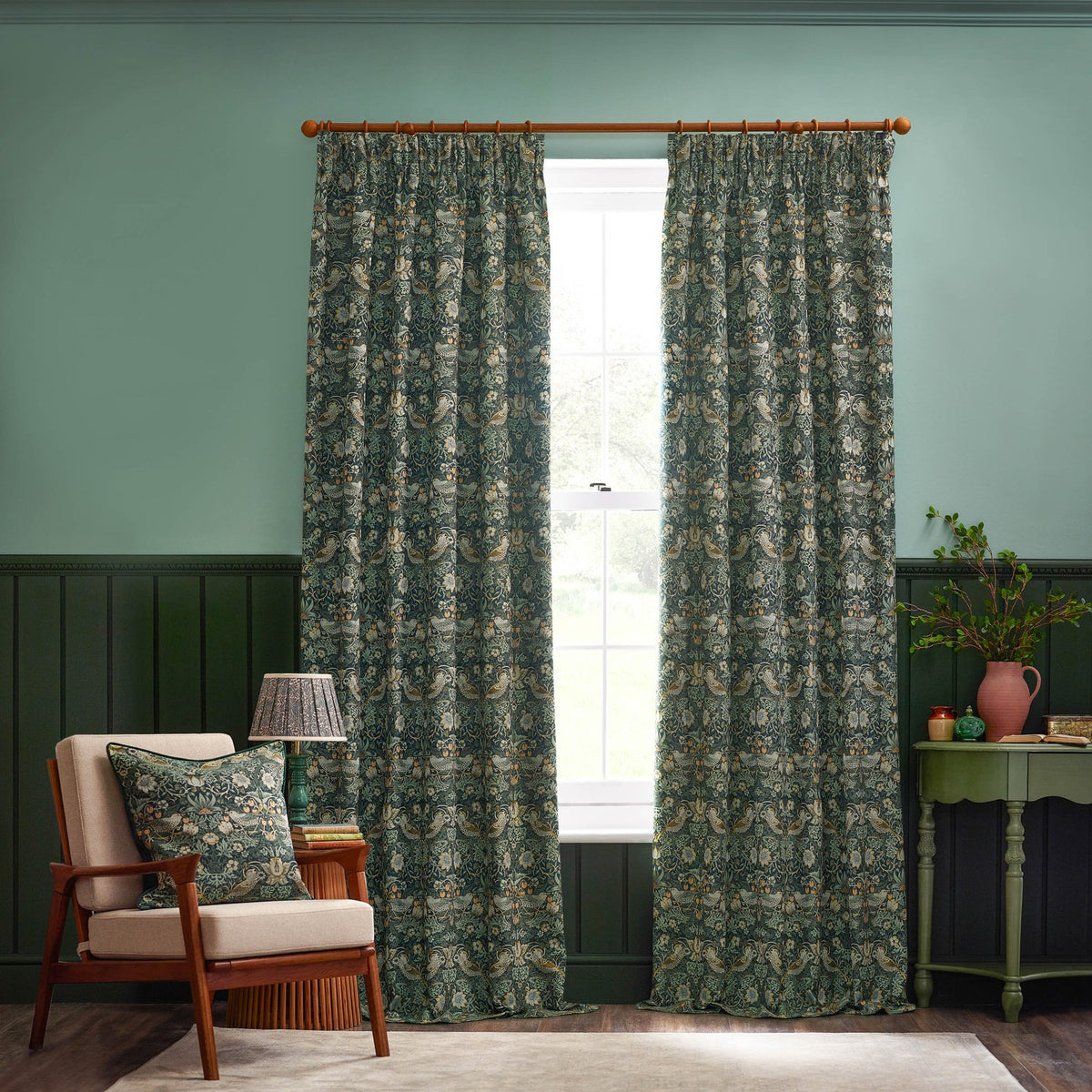 Strawberry Thief Jacquard Velvet Cushion in Forest Green on a beige armchair, showing woven bird and foliage pattern in cream, gold and amber tones, in a living room setting.