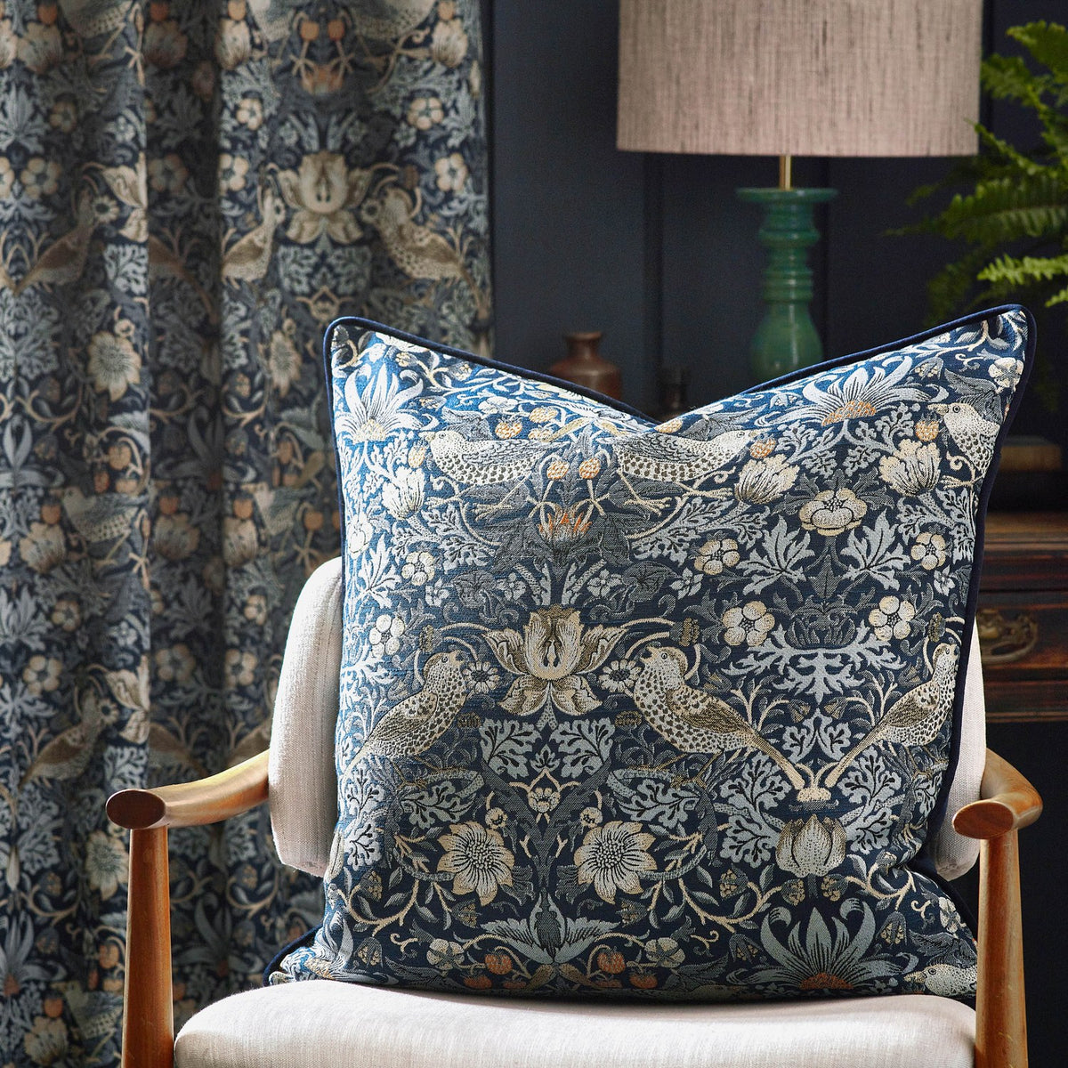 Strawberry Thief Jacquard Velvet Cushion in indigo with bird and foliage pattern, navy piped edge, styled on a light upholstered armchair against patterned curtains.
