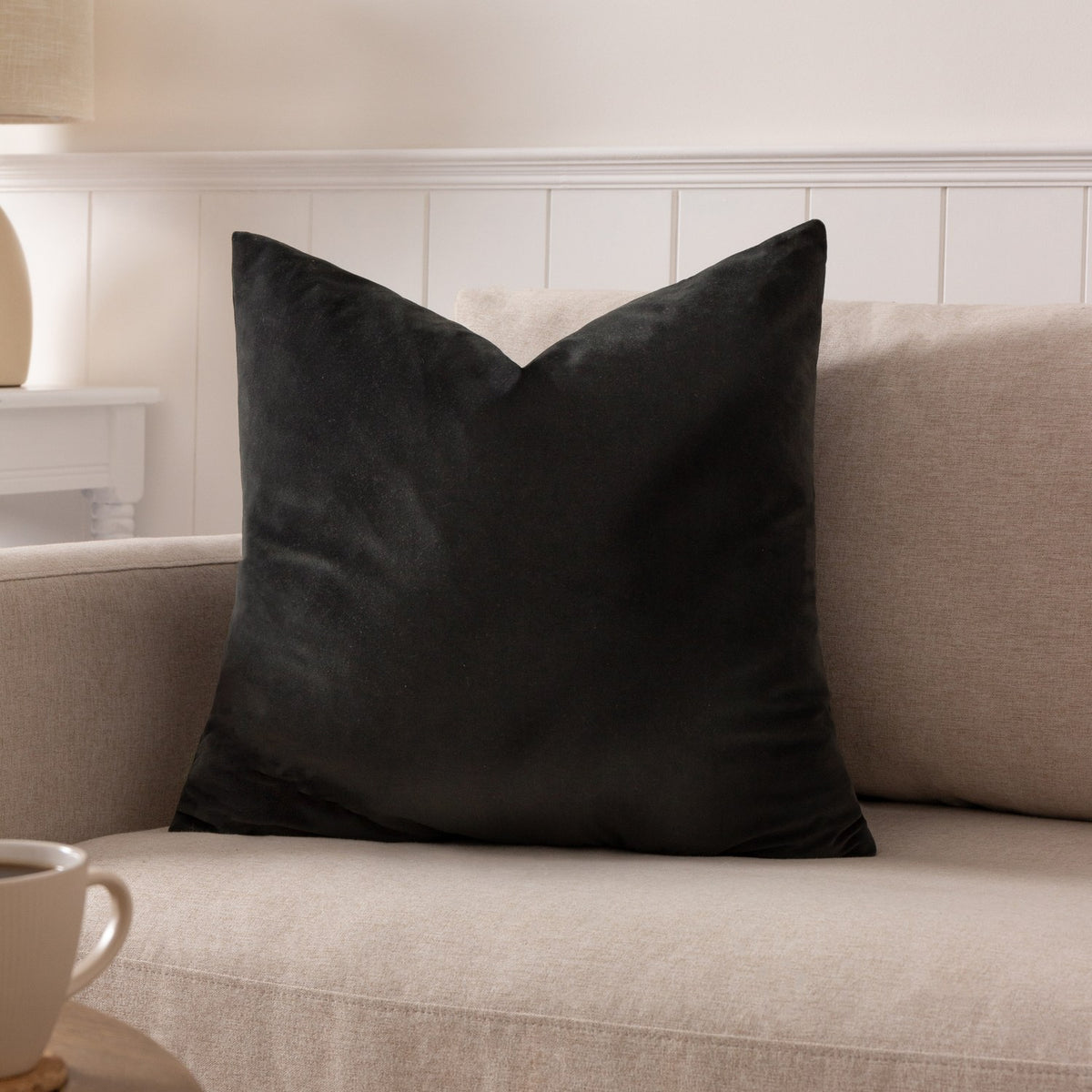 Sunningdale Plain Velvet Cushion in charcoal, square with pointed corners, shown on a beige sofa in a living room setting.