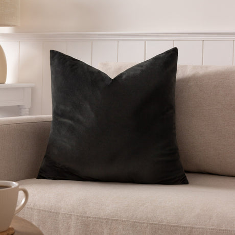 Sunningdale Plain Velvet Cushion in charcoal, square with pointed corners, shown on a beige sofa in a living room setting.
