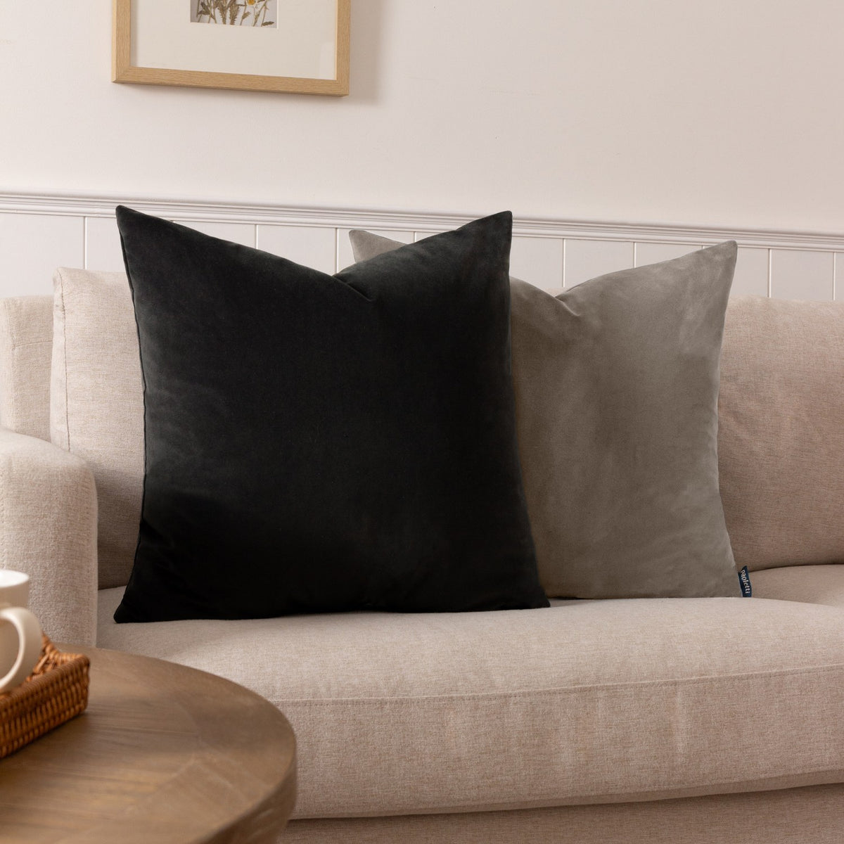 Sunningdale square velvet cushion in charcoal grey, shown on a beige sofa beside a black velvet cushion in a living room setting.