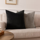 Sunningdale square velvet cushion in charcoal grey, shown on a beige sofa beside a black velvet cushion in a living room setting.