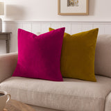 Sunningdale plain velvet cushion in deep cerise pink, shown square with a mustard velvet cushion on a beige sofa in a living room setting.