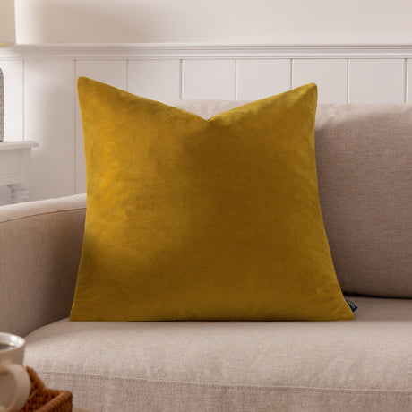 Sunningdale Plain Velvet Cushion in saffron gold, square polyester velvet cushion shown on a beige sofa in a living room setting.
