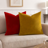 Sunningdale plain velvet square cushion in saffron gold, shown on a beige sofa beside a red velvet cushion in a living room setting.
