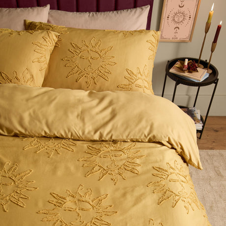 Gold duvet cover set with tufted sun motifs on pillowcases and duvet, shown on a bed in a bedroom with candles and a side table.