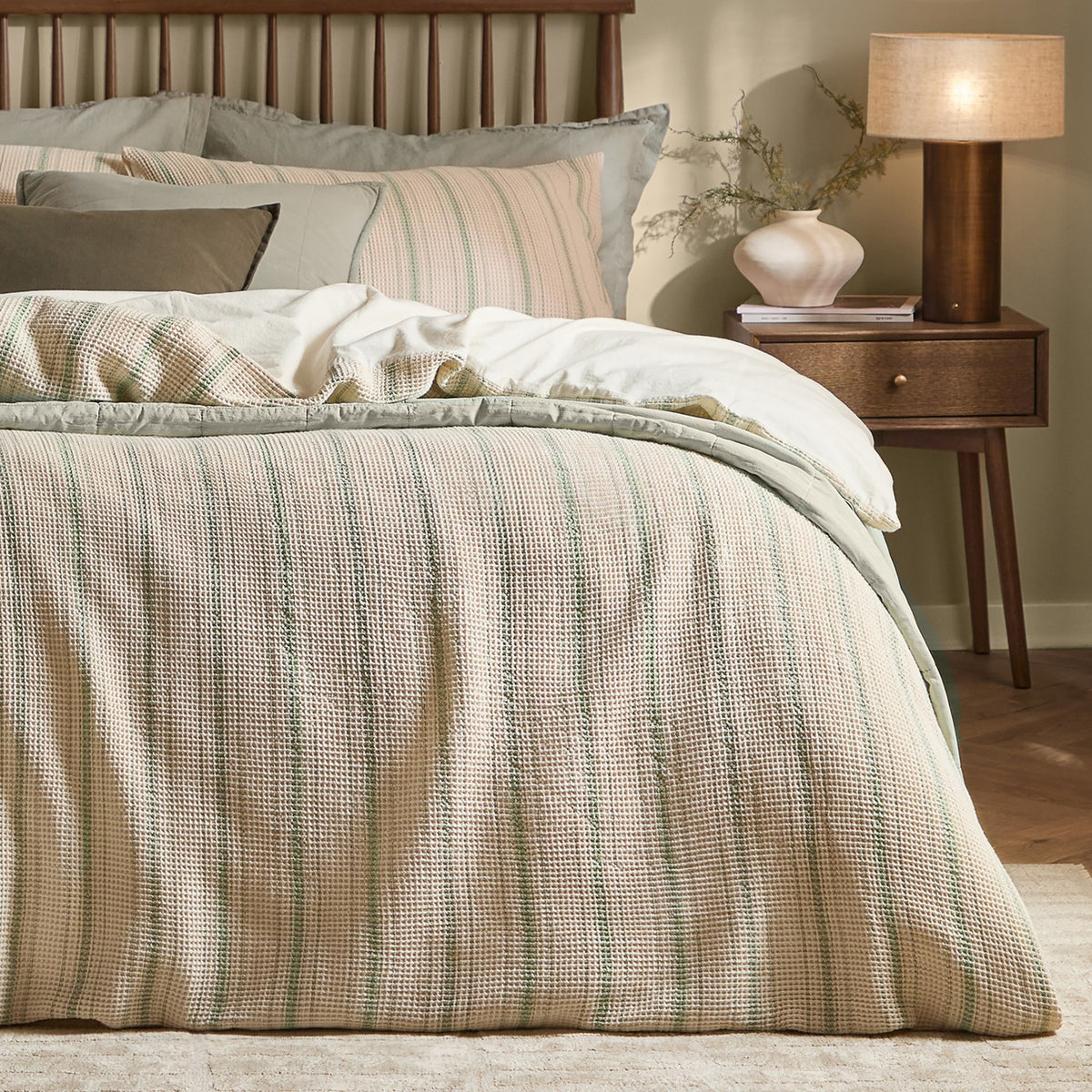 Selby Waffle Cotton Striped Duvet Cover Set in bay green and natural on a bed with layered cushions, styled beside a wooden nightstand with lamp in a bedroom setting.