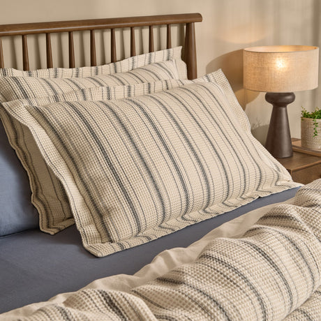 Selby Waffle Percale Cotton Pillowcase Pair in coal and natural, shown stacked on a bed with charcoal striped waffle texture, styled with a wooden headboard and bedside lamp.