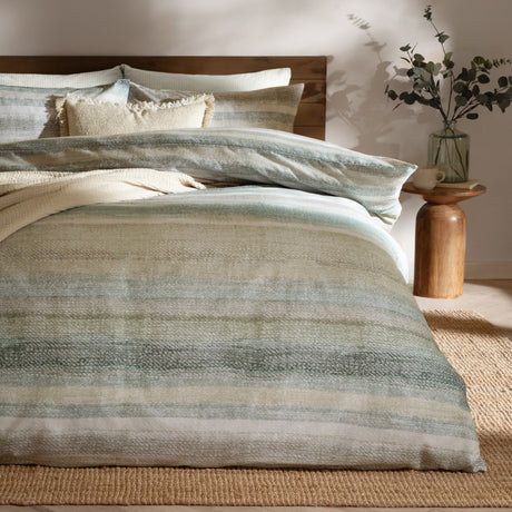 Swift Cotton Striped Duvet Cover Set in Eucalyptus, dressed on a bed with matching pillowcases, cushions and a fringed cushion, showing muted green, sage and sand stripes in a bedroom setting.
