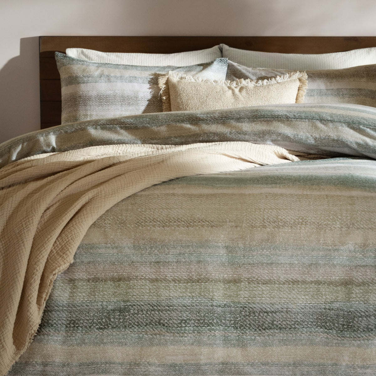 Swift Cotton Striped Duvet Cover Set in Eucalyptus, styled on a bed with matching pillowcases in muted green tones, layered with cream cushions and a beige throw against a wooden headboard.
