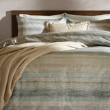 Swift Cotton Striped Duvet Cover Set in Eucalyptus, styled on a bed with matching pillowcases in muted green tones, layered with cream cushions and a beige throw against a wooden headboard.