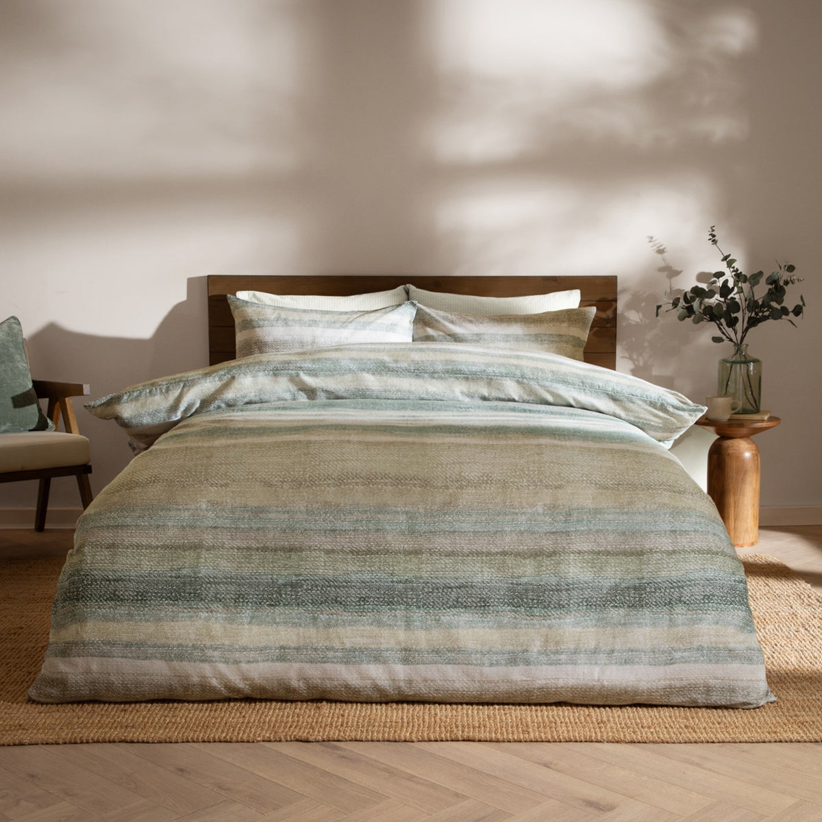 Swift Cotton Striped Duvet Cover Set in Eucalyptus, styled on a bed with matching pillowcases in muted green, sage and sand stripes, set in a bedroom with wooden headboard and side table.