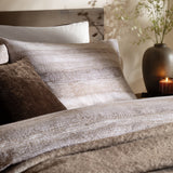 Swift Cotton Striped Duvet Cover Set in natural tones, styled on a bed with matching pillows, a brown cushion and a bedside vase with candle.