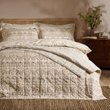 Quilted Strawberry Thief cotton bedspread in natural beige tones on a bed with striped pillowcases and shams, in a bedroom with wooden headboard.