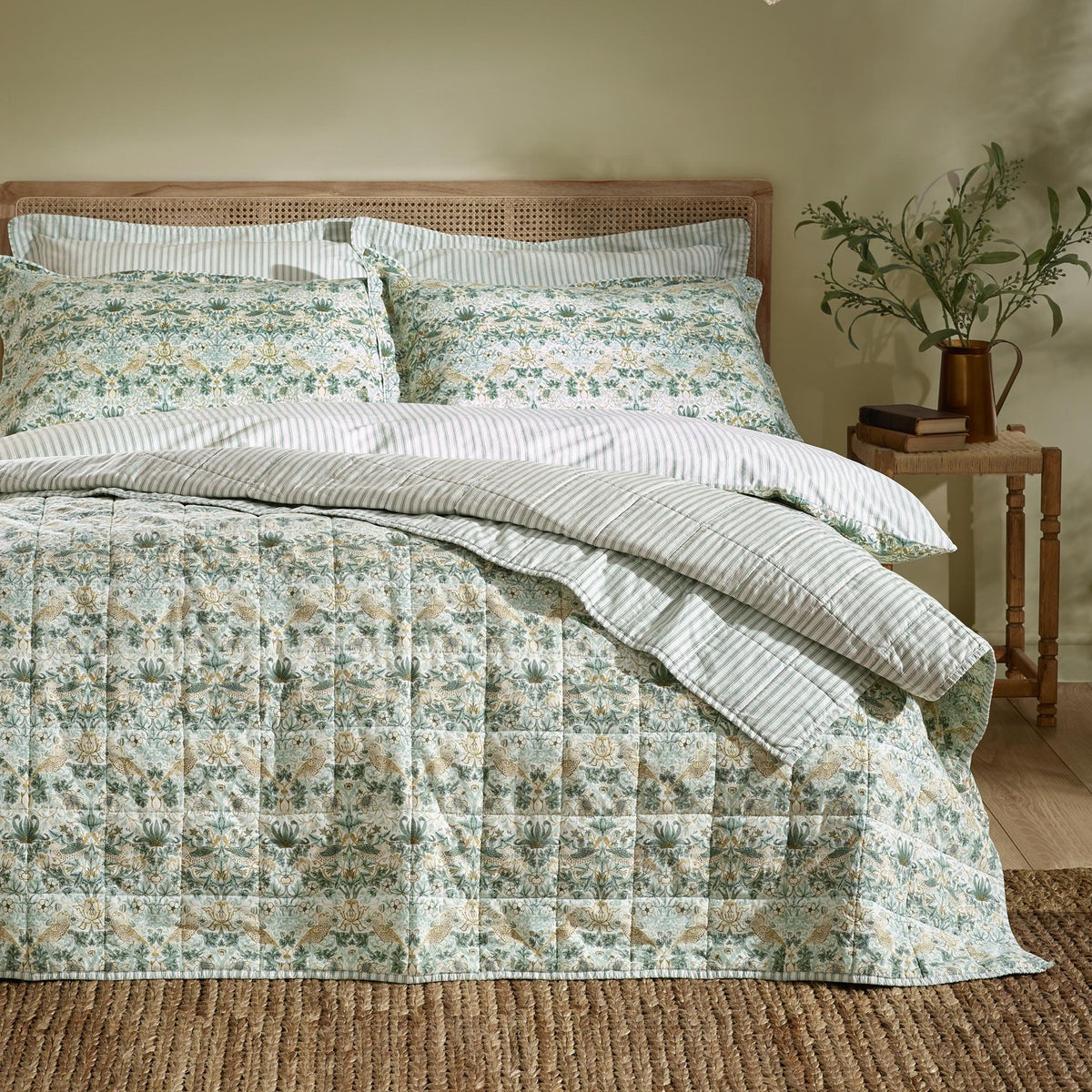 Strawberry Thief quilted cotton bedspread in sage green with floral bird and berry print, shown on a bed with striped pillowcases and a rattan headboard in a bedroom setting.