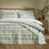 Strawberry Thief quilted cotton bedspread in sage green with floral bird and berry print, shown on a bed with striped pillowcases and a rattan headboard in a bedroom setting.
